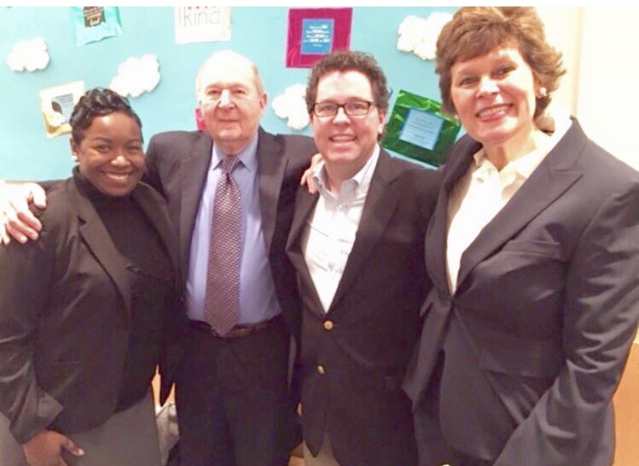 Four people in business attire posing together beside a colorful bulletin board
