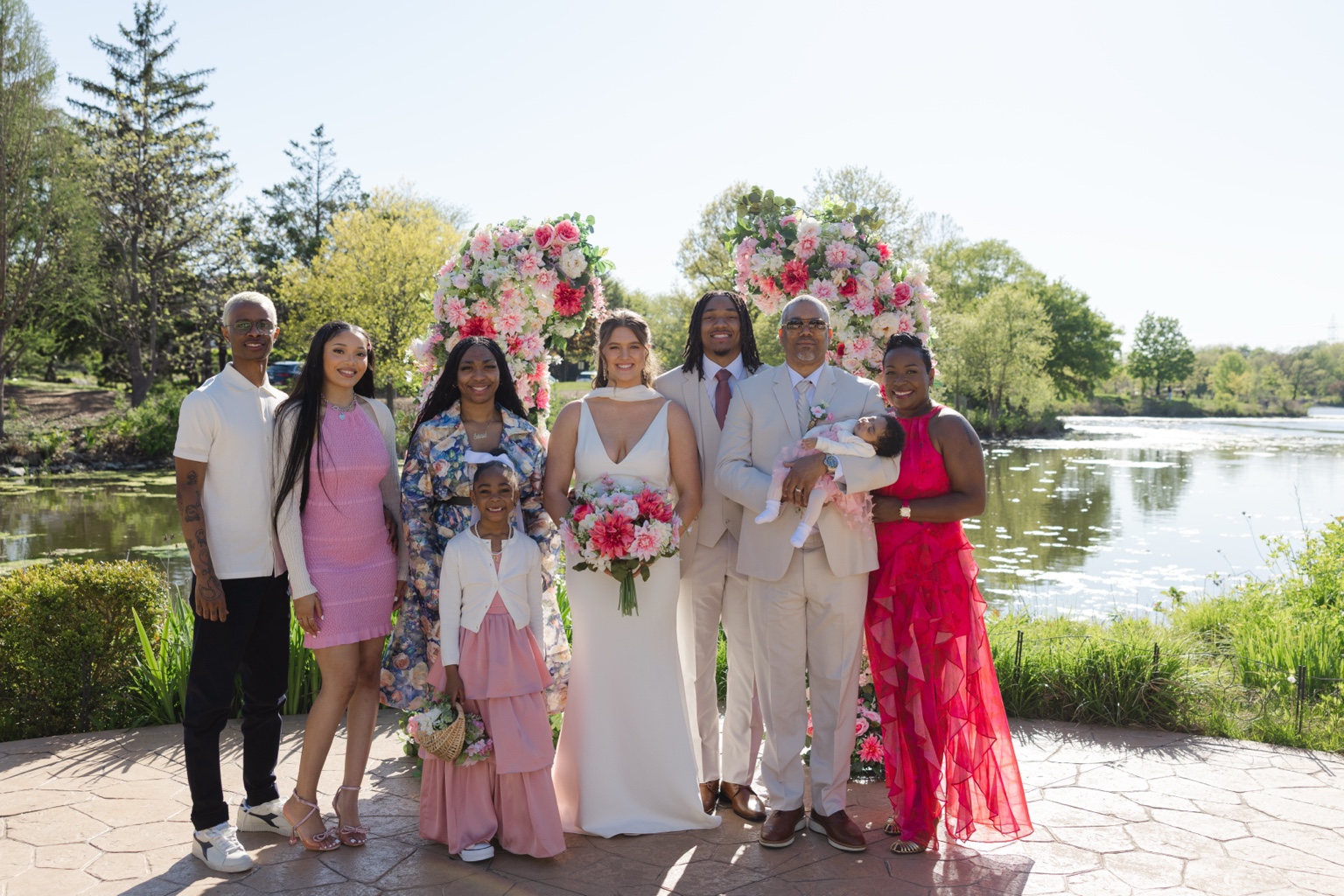 Wedding group portrait with adults and children beside a lake and pink floral arches