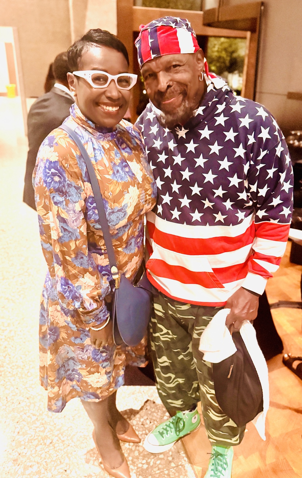 Two people posing together, one in a floral dress and white glasses and the other in stars and stripes