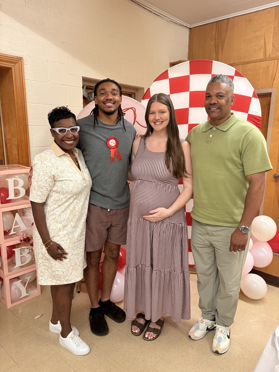 Four people celebrating at a baby shower with balloons and a red-and-white backdrop