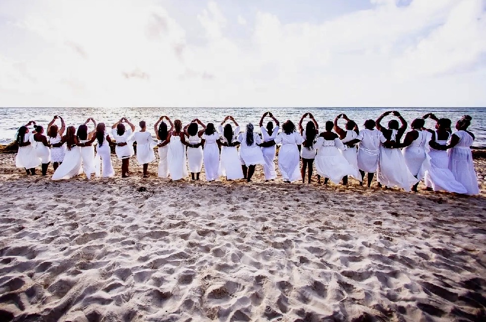 A group dressed in white standing arm in arm on a beach facing the water