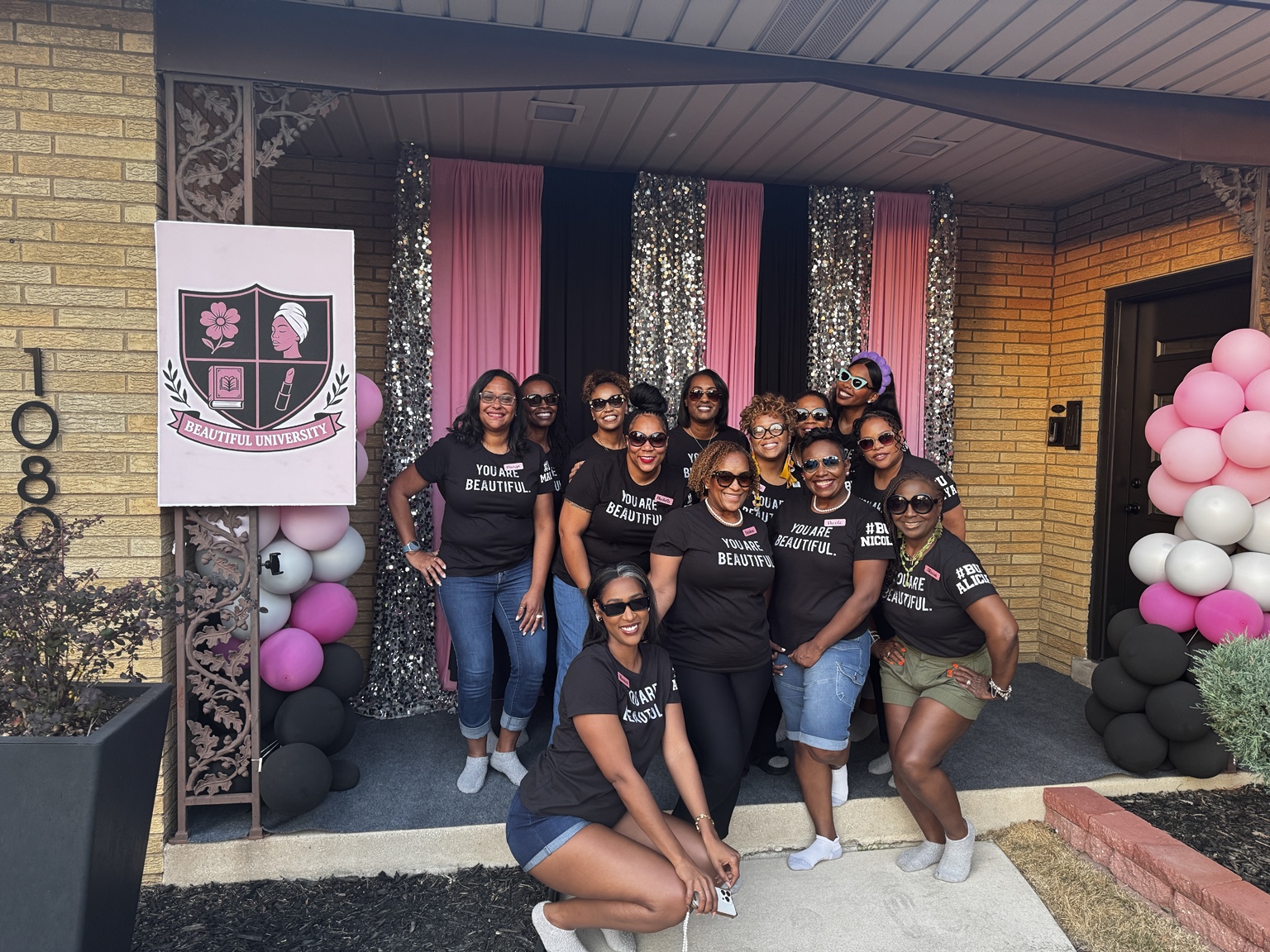 A group in matching You Are Beautiful shirts posing beside pink balloons and a Beautiful University sign