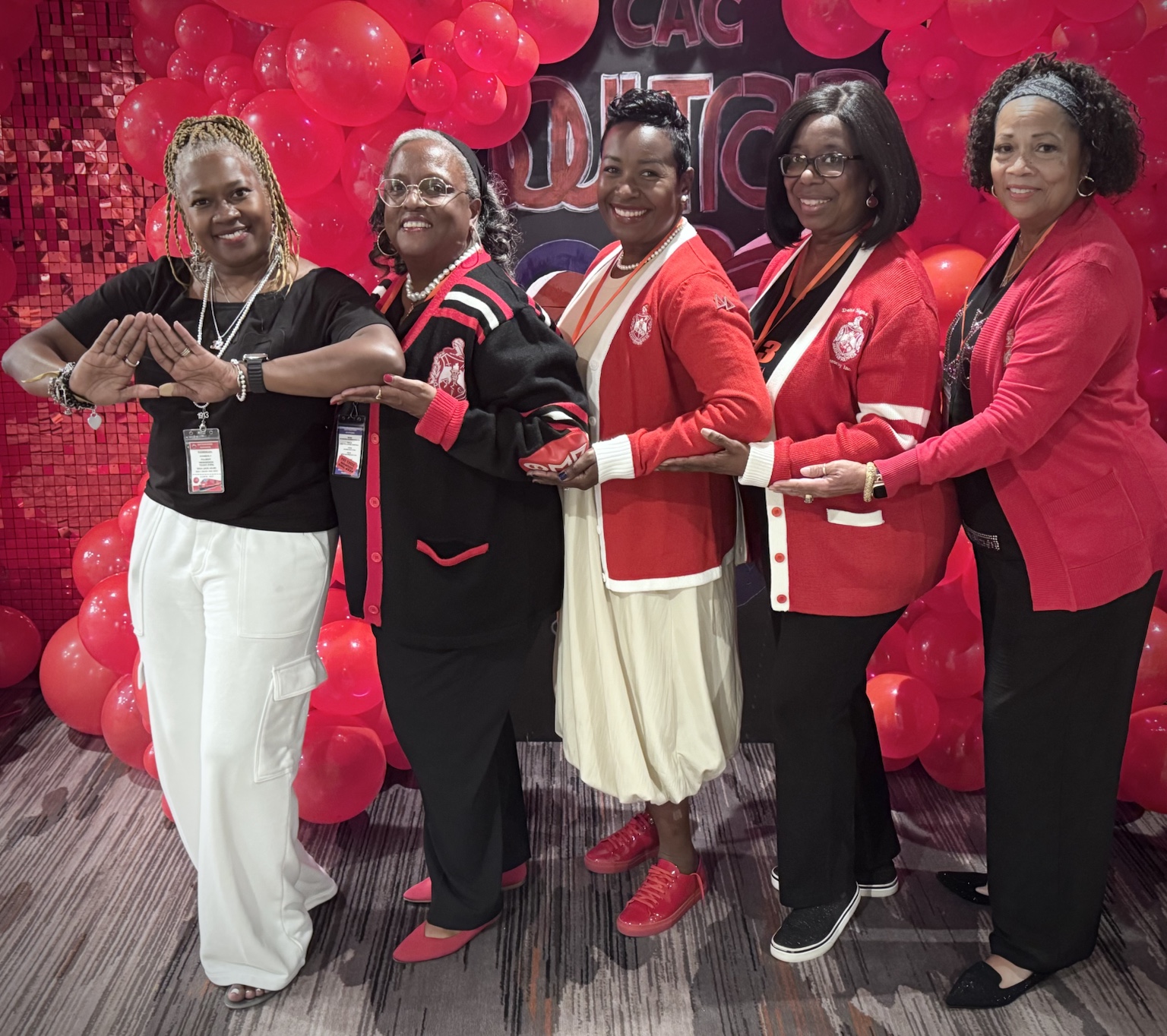 Five women in red, white, and black sorority apparel posing in front of red balloons